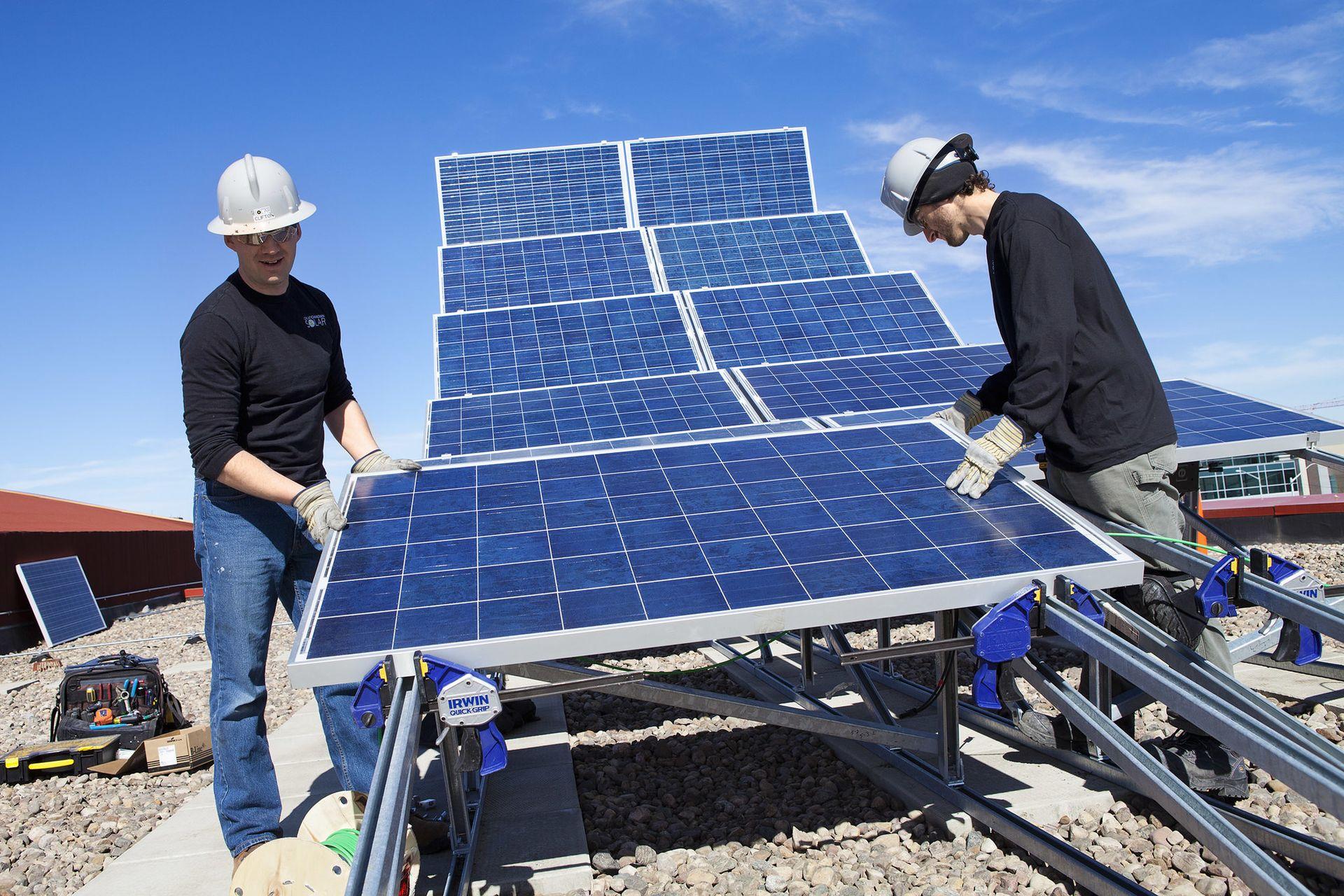 Two workers in hardhats installing solar panels on a rooftop under a clear blue sky, with tools and equipment nearby.